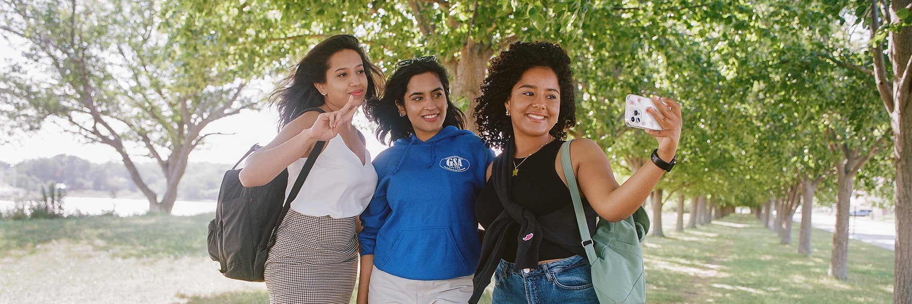 3 students take selfie under trees.