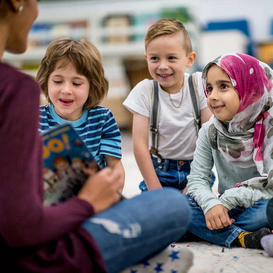 A multi-ethnic group of young school children are indoors in their classroom. Their teacher is reading a storybook to them.