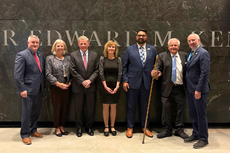 People stand side by side in formal and semi-formal attire, smiling in front of a dark stone wall.