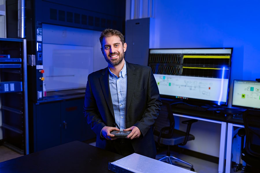 Ioannis Zografopoulos smiles at the camera, surrounded by the equipment he uses to conduct his research. A futuristic blue light glows in the background.