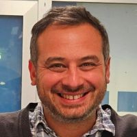 Man with a big smile, short hair, a plaid button shirt, and a brown seater.