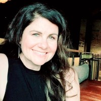 Photo of woman with long, dark hair. She is wearing a black top and is facing forward with a big smile.